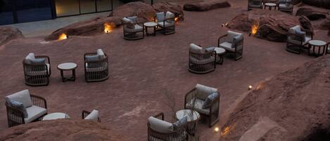 An outdoor seating area with wicker furniture and tables, surrounded by large rocks and a building in the background.
