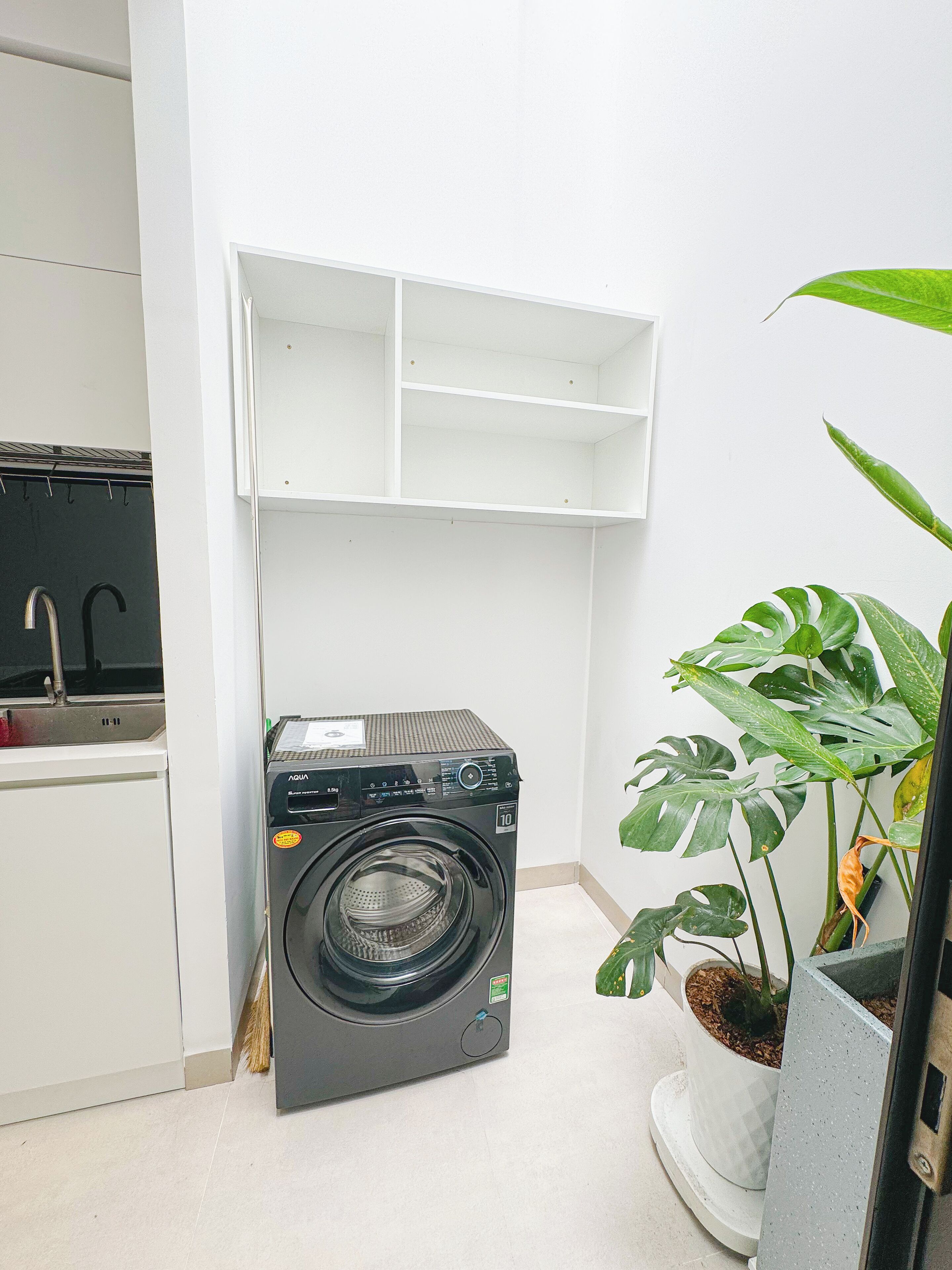 A compact laundry area with a front-loading washing machine, a sink, and a white shelving unit.