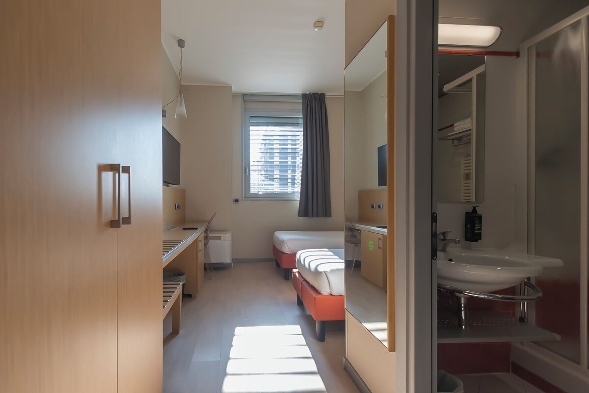 A hotel room with a bed, a desk, a window with blinds, a bathroom with a sink and mirror, and a wardrobe.