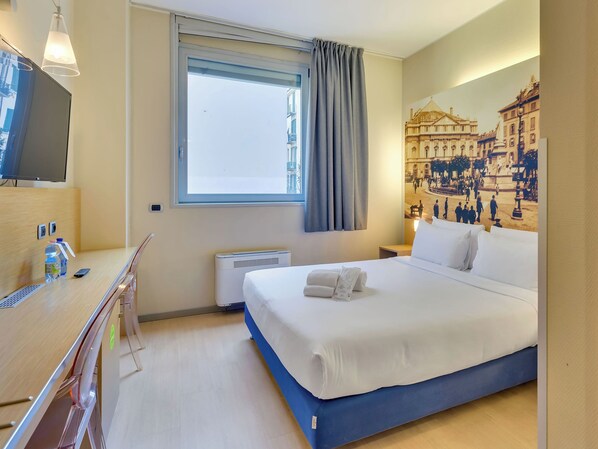 Hotellihuone, jossa on sänky, työpöytä, televisio, ikkuna verhoilla ja seinämaalaus.