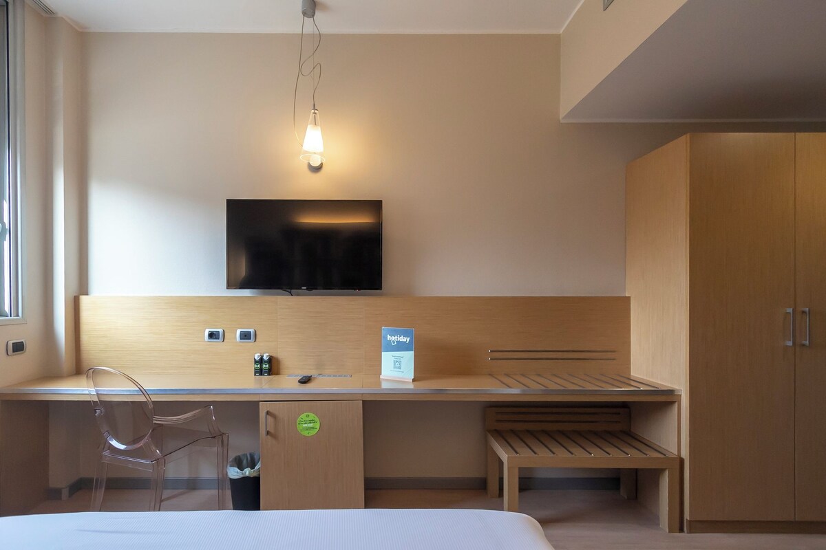 A hotel room with a bed, a desk, a chair, a television, and a wardrobe.