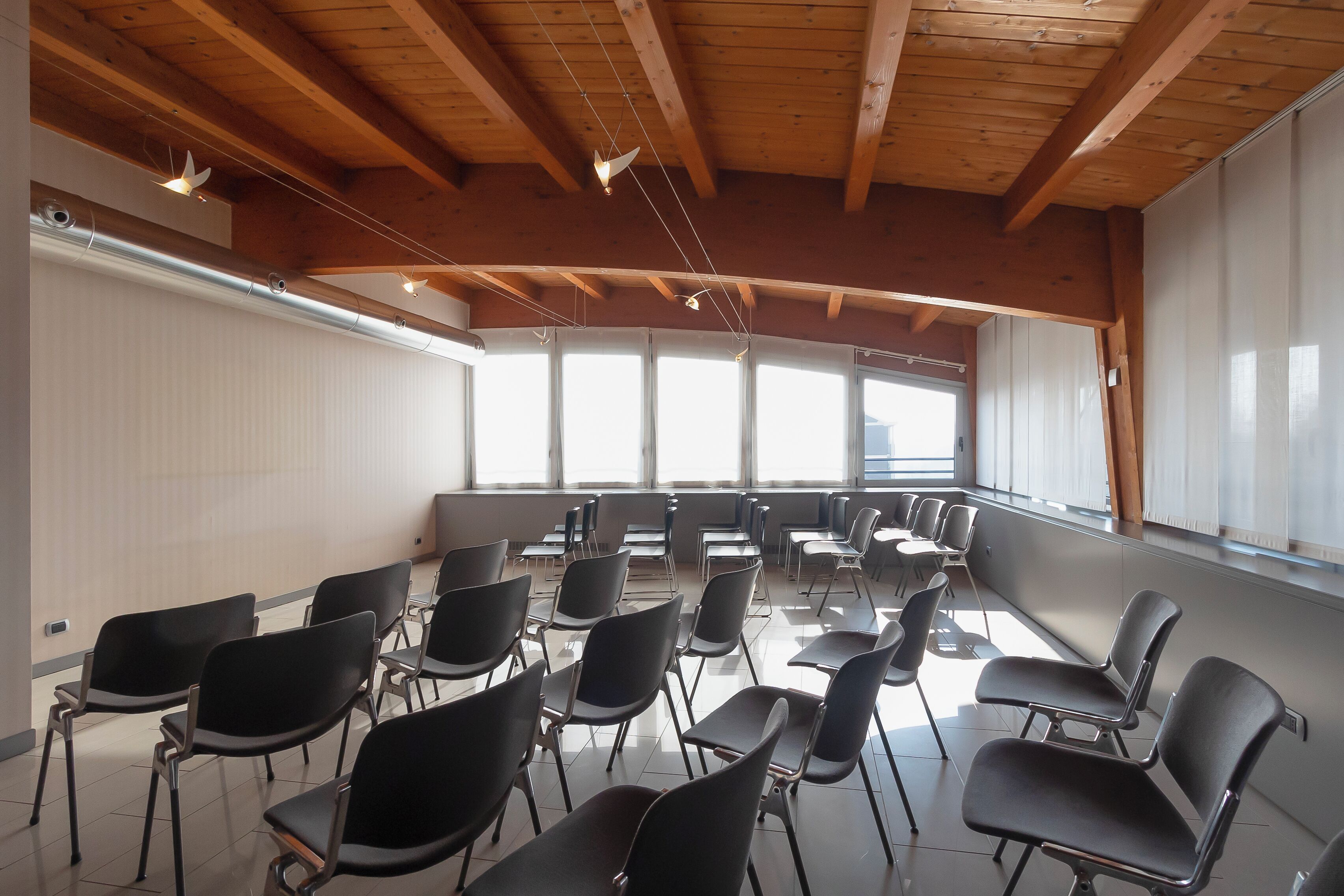 Un salón de conferencias con hileras de sillas, vigas de madera en el techo y grandes ventanales.