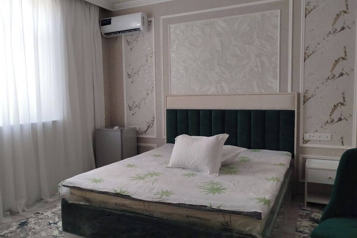 Una habitación de hotel con una cama, una mesita de noche, una silla verde y una ventana con cortinas traslúcidas.