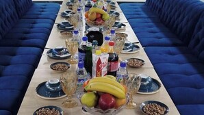 A long dining table with blue upholstered sofas on either side, set with plates, bowls, glasses, and bottles. - SUNBULA (Denov)