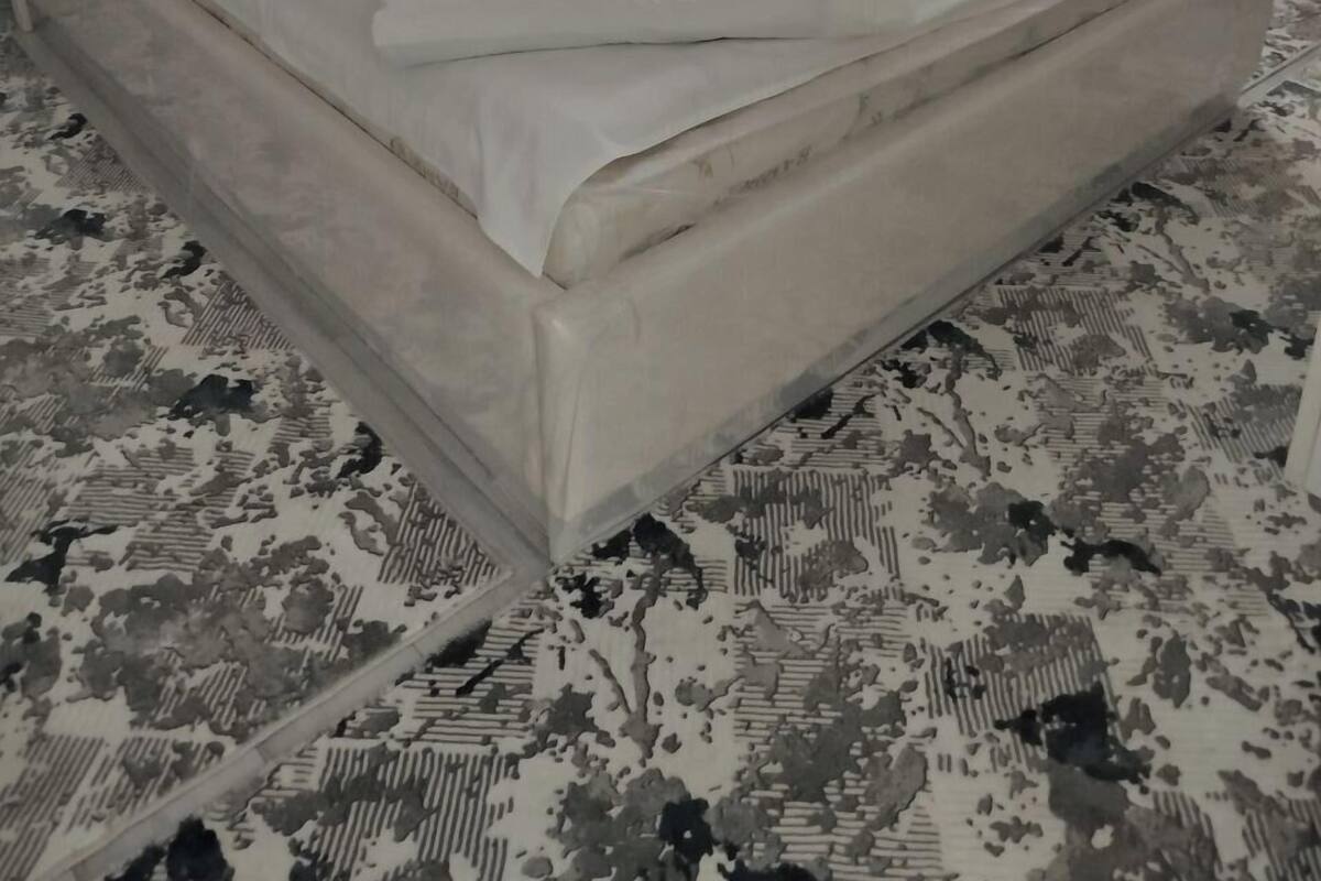 Una cama bien hecha con cabecera blanca y almohadas, una alfombra estampada y una ventana con cortinas de encaje.