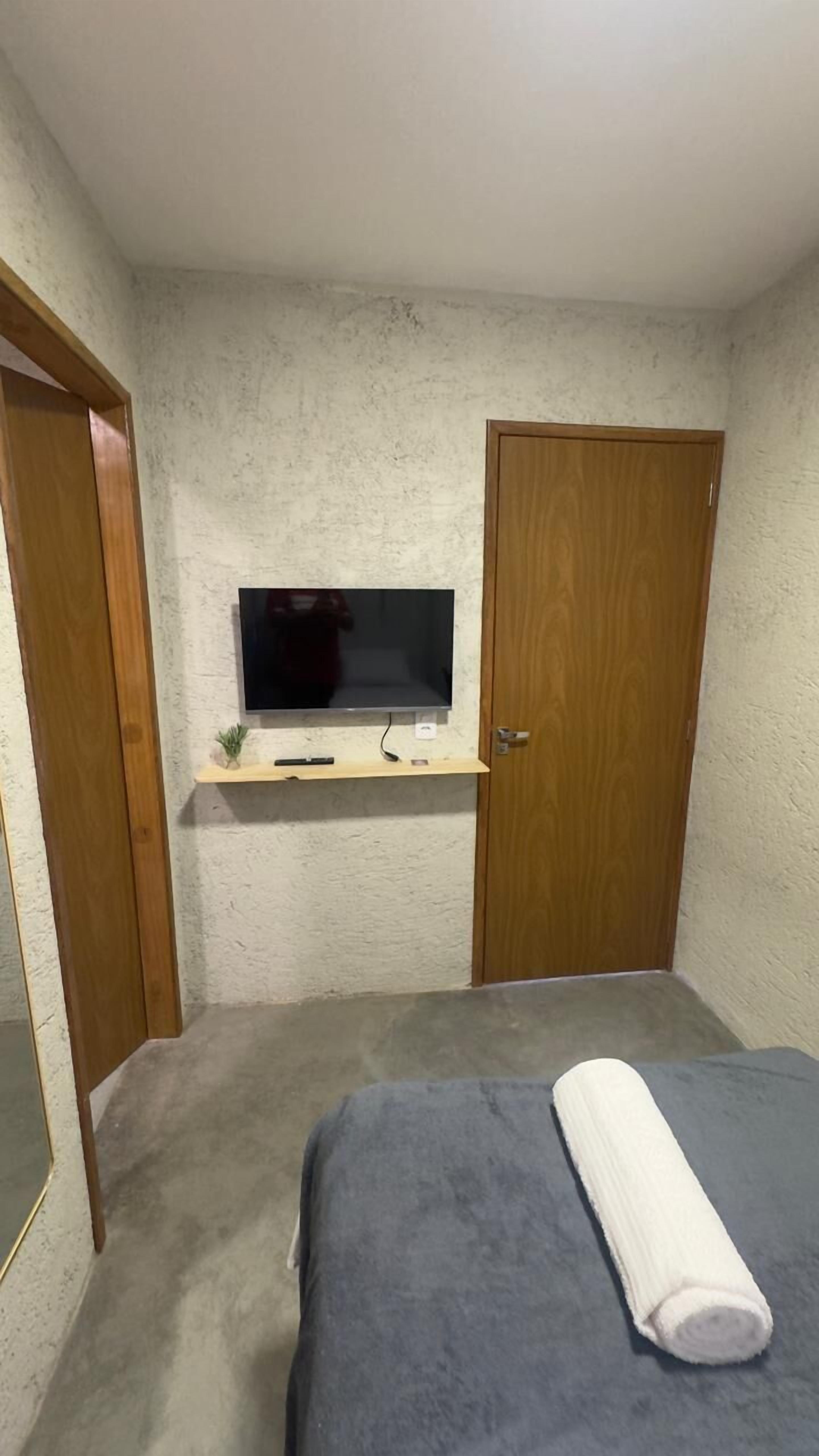 Quarto de hotel pequeno e individual, com uma cama, uma televisão fixada na parede e uma porta de madeira.