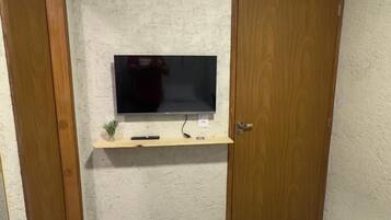 Quarto de hotel pequeno e individual, com uma cama, uma televisão fixada na parede e uma porta de madeira.