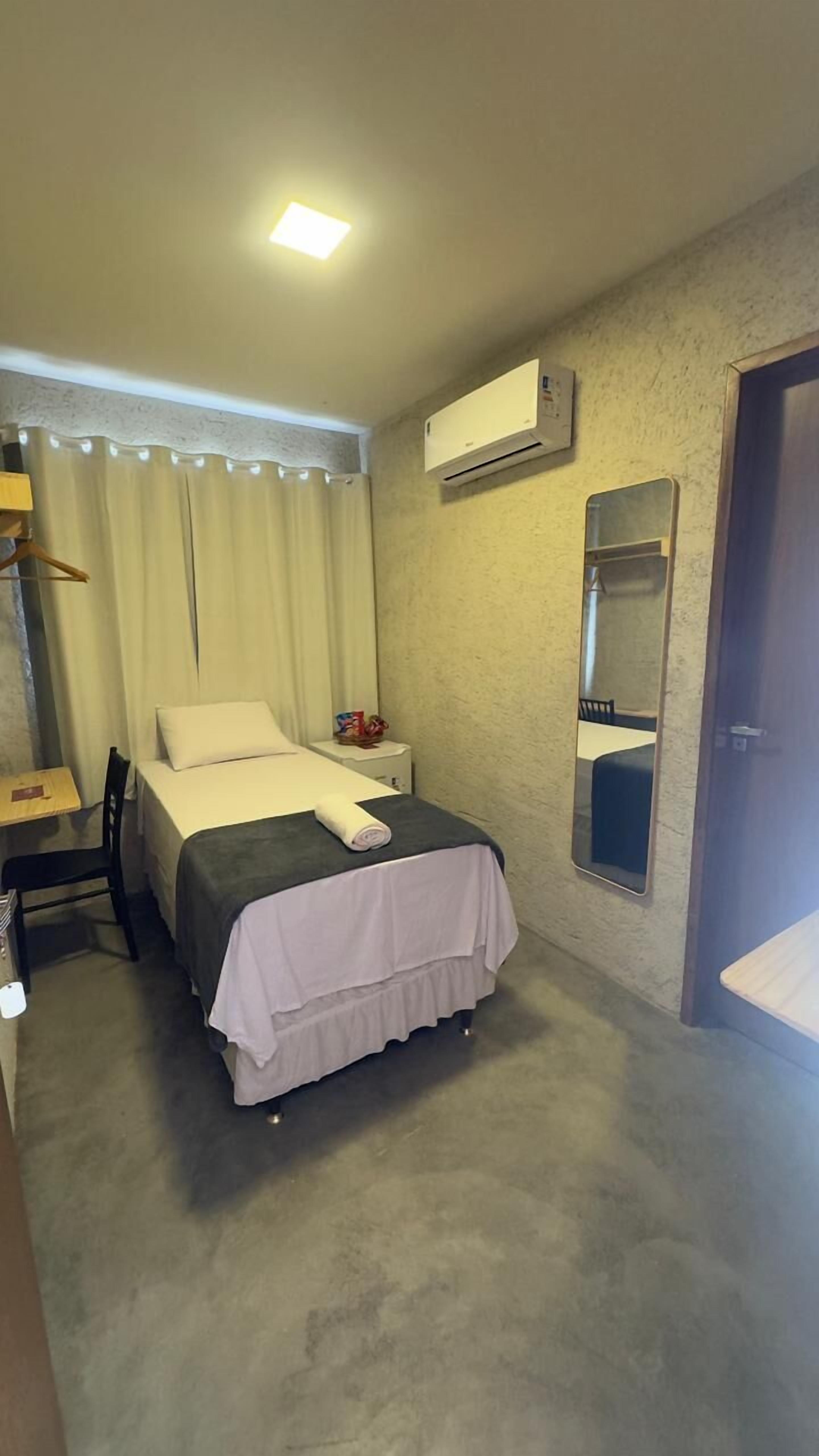 A small, neatly arranged hotel room with a single bed, a bedside table, a chair, a desk, and a wardrobe.