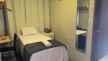 A small, neatly arranged hotel room with a single bed, a bedside table, a chair, a desk, and a wardrobe.