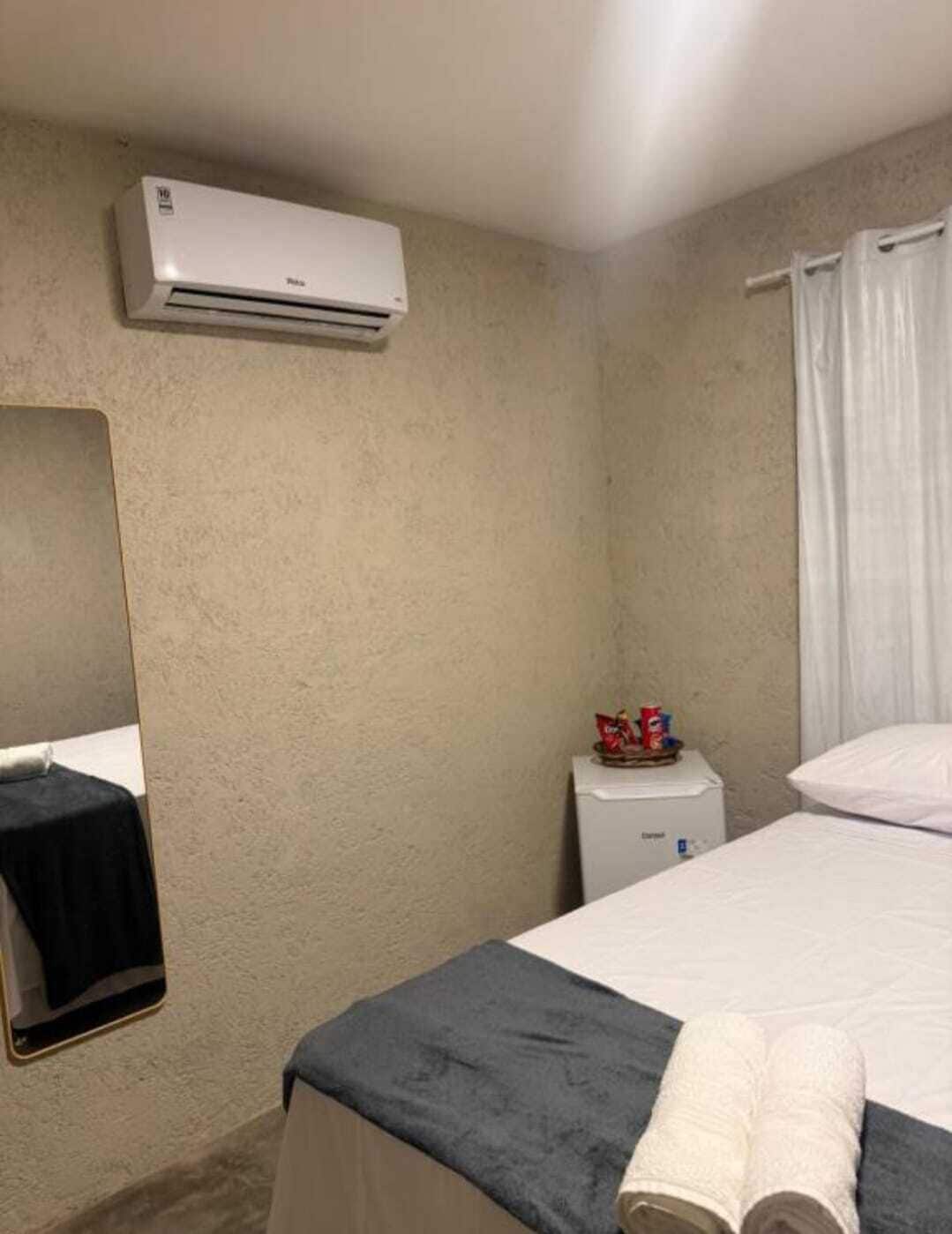Quarto de hotel com cama, uma pequena mesa de cabeceira com um objeto vermelho, ar condicionado na parede, um espelho e uma janela com cortinas brancas.
