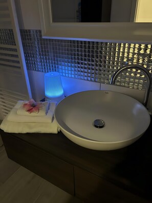 Ein Badezimmer mit einem runden Waschbecken, einem Handtuch, einer Blume und einer blau beleuchteten Lampe.