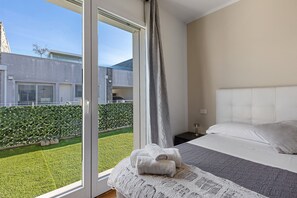 Een slaapkamer met een bed, een raam met gordijnen en uitzicht op een groen gazon en een gebouw.