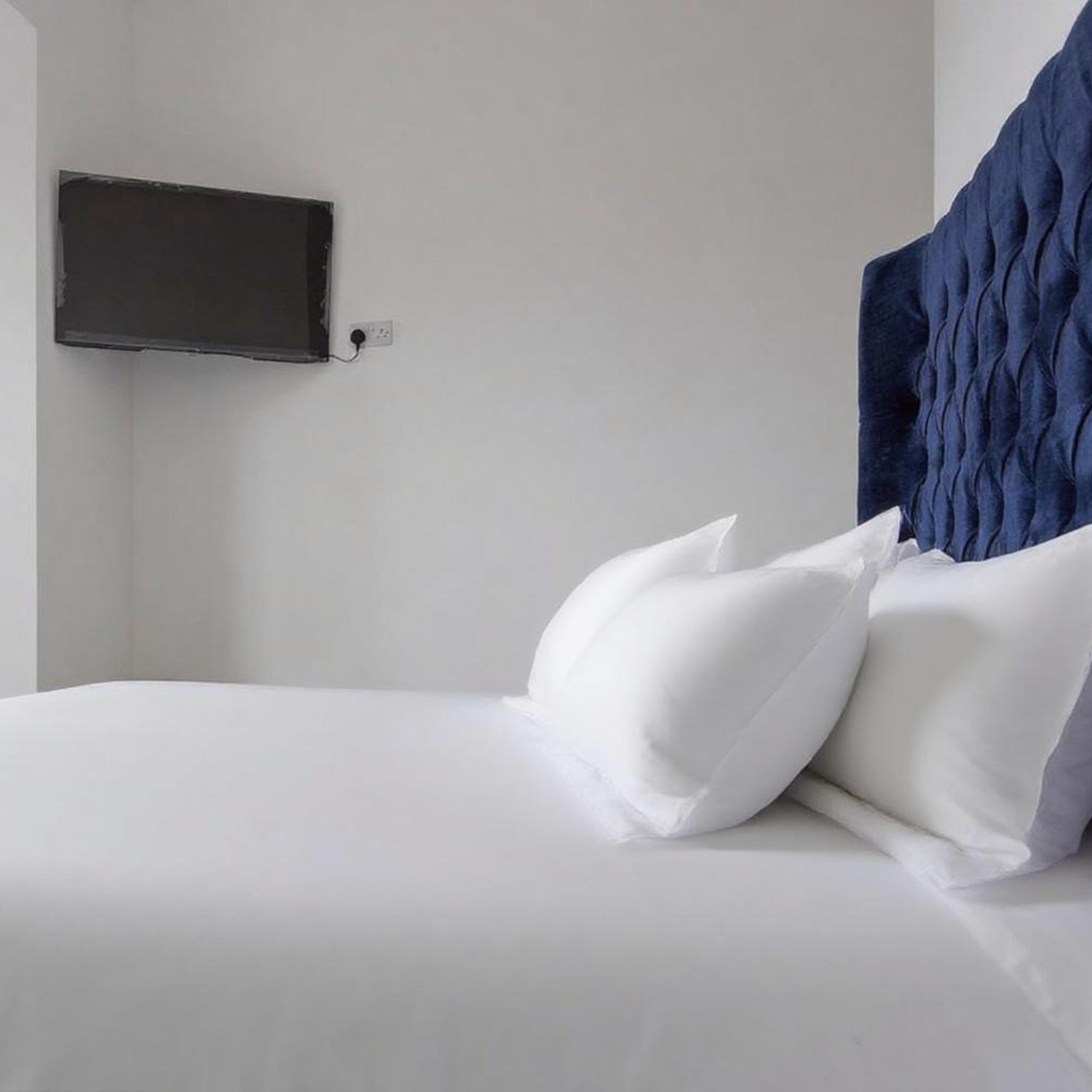 A neatly made bed with white linens and two white pillows, a dark blue headboard, and a flat-screen TV mounted on the wall.