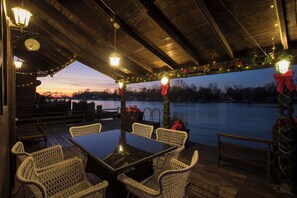 A covered outdoor deck with a table and chairs, decorated with lights and greenery, overlooking a body of water.