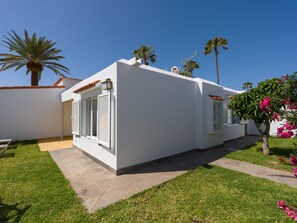 Een wit gebouw met een betegeld dak, een kleine tuin met gras en een boom, en een heldere blauwe lucht.