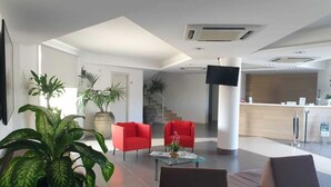 Een moderne lobby met een receptiebalie, rode stoelen, een bank en potplanten.