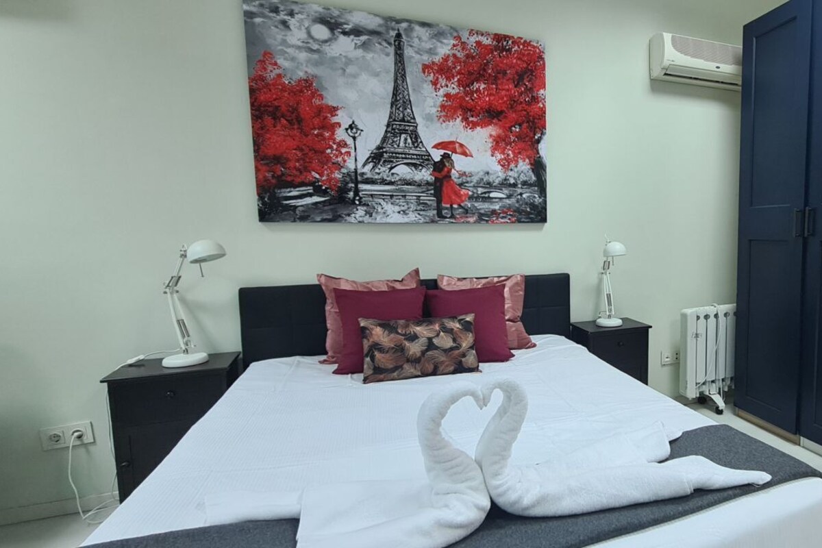 A bedroom with a bed, pillows, a nightstand, a lamp, an air conditioner, and a door.