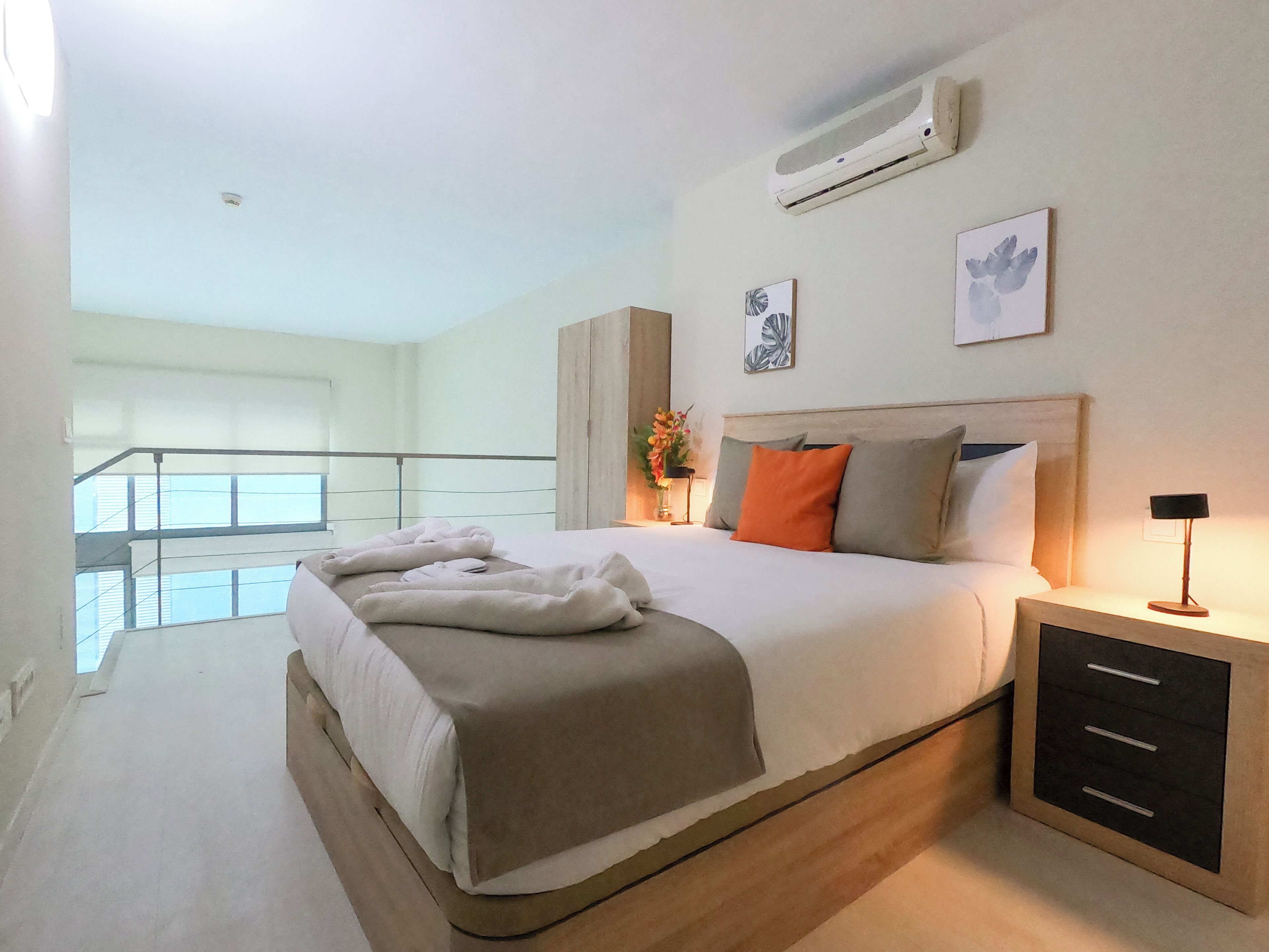 A modern hotel room with a large bed, a nightstand, a wall-mounted air conditioner, and a balcony with a railing.