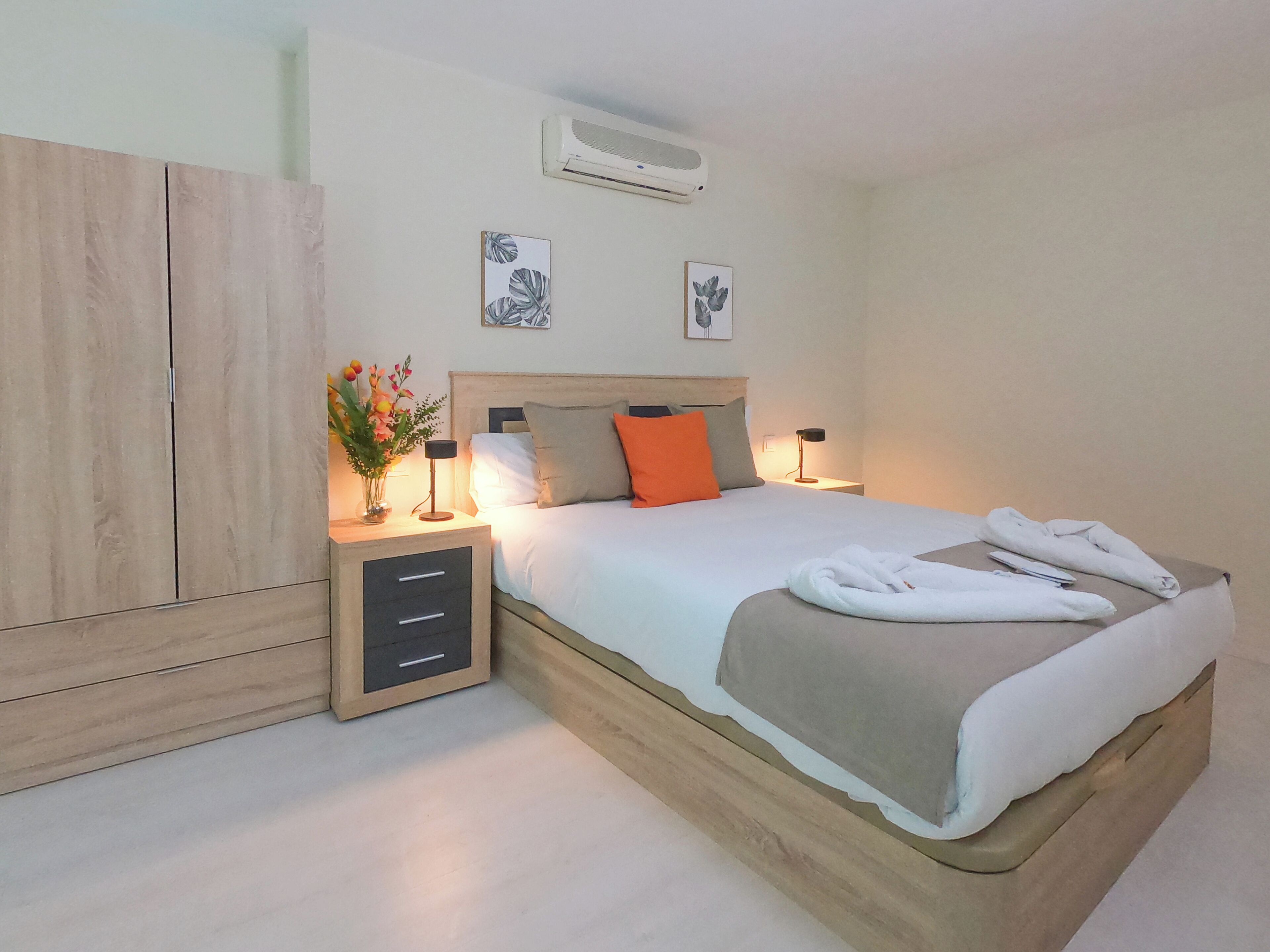 A neatly arranged bedroom with a bed, bedside lamps, a nightstand, a wardrobe, and a wall-mounted air conditioner.