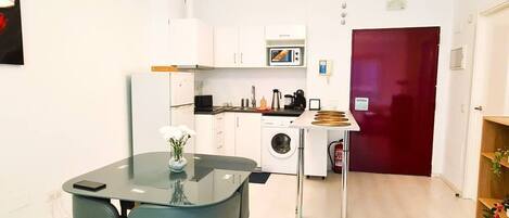 A compact kitchen with a microwave, sink, and washing machine. There is a glass dining table with chairs and a vase of flowers.