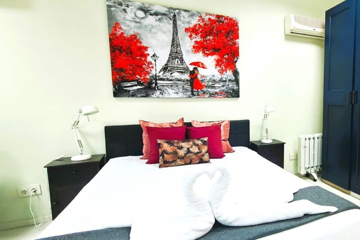 A neatly made bed with white linens and a decorative pillow, a black nightstand with a lamp, and a large painting of the Eiffel Tower.