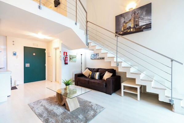 A modern living room with a glass coffee table, a brown leather sofa, and a staircase with white steps. - Oshun Sanchinarro Seasonal Living (Madrid)