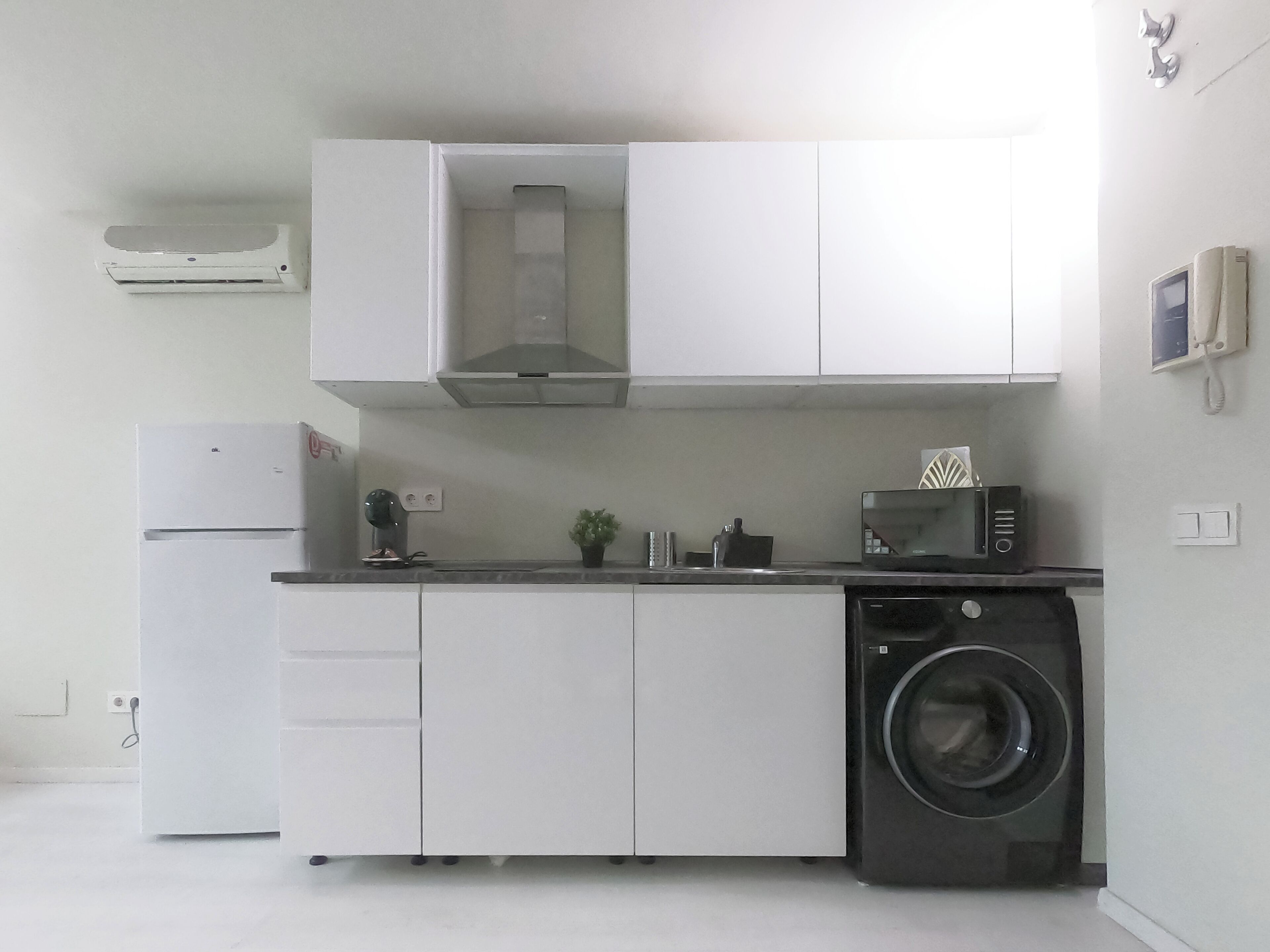 Una cocina compacta con armarios blancos, microondas integrado, lavadora y cafetera.