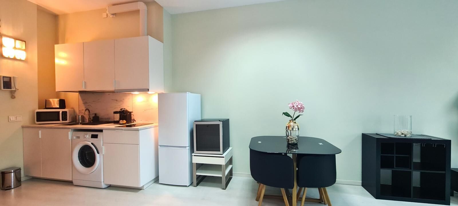 A compact kitchen with a washing machine, microwave, and a small refrigerator. There is a black table with a vase and a potted plant.