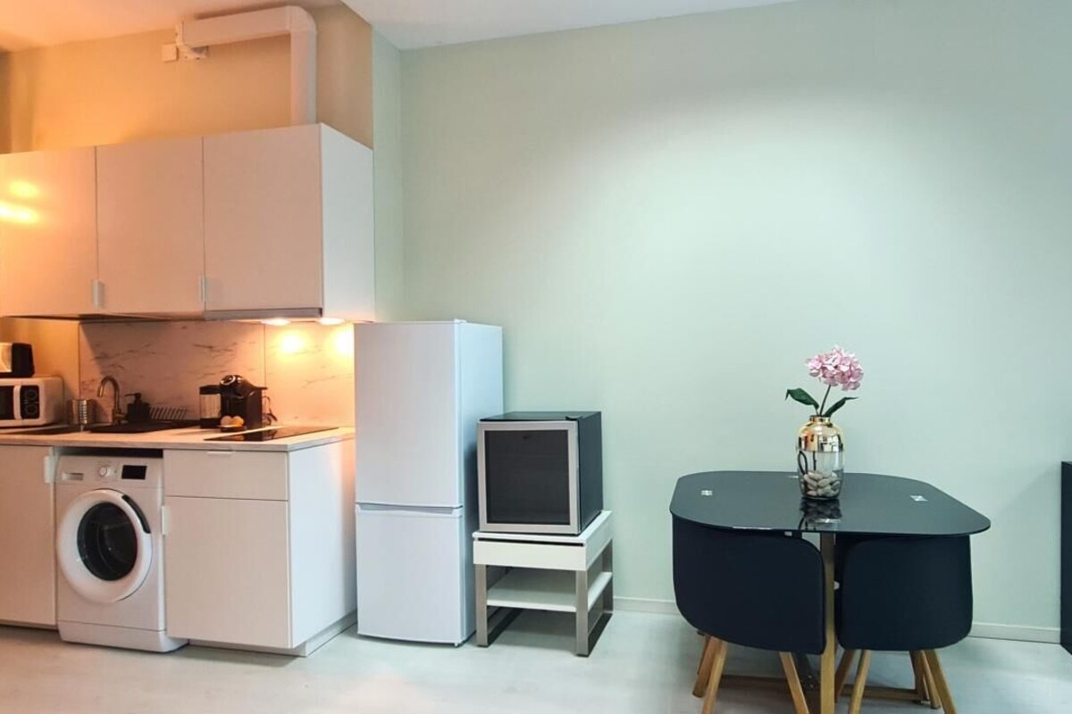 A compact kitchen with a washing machine, microwave, and a small refrigerator. There is a black table with a vase and a potted plant.