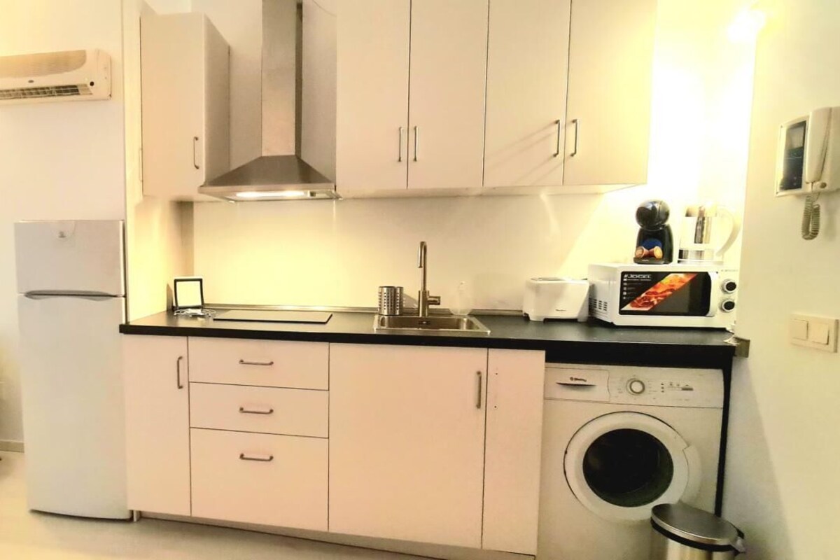 Eine kompakte Küche mit Kühlschrank, Mikrowelle, Spüle und Waschmaschine. Ein Esstisch mit Stühlen und eine kleine Pflanze sind ebenfalls vorhanden.