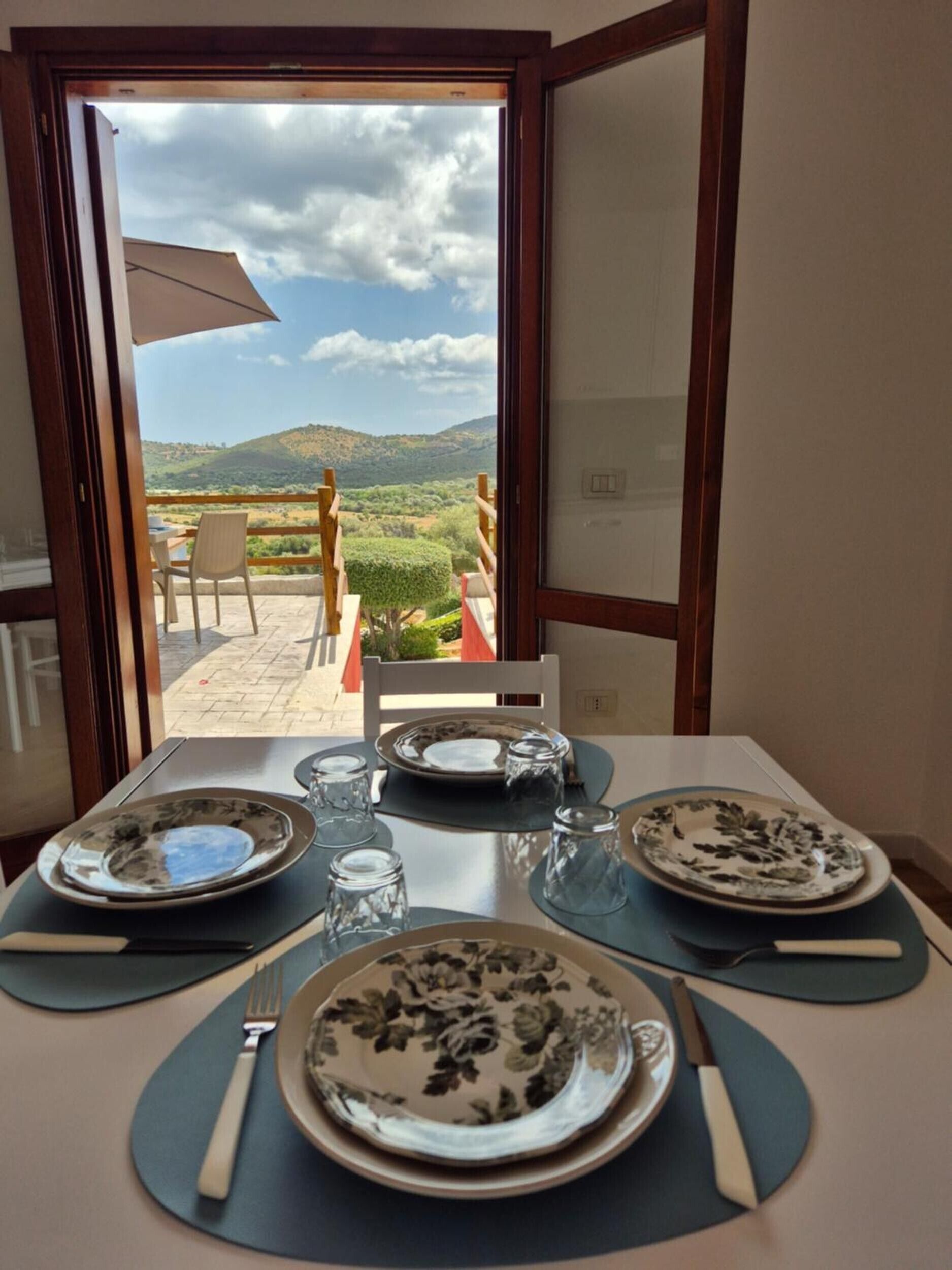 A dining table set with plates, cutlery, and glasses, with a view of a mountainous landscape through an open door.