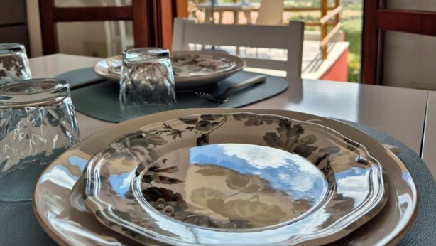 Une table à manger dressée avec des assiettes, des verres et des couverts, donnant sur une terrasse et un ciel nuageux.