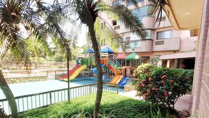 Un parque infantil con toboganes y estructuras para escalar, rodeado de follaje y una piscina.