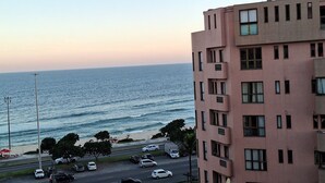 Escena urbana costera con un edificio de varios pisos, una calle con coches estacionados y una playa al fondo.