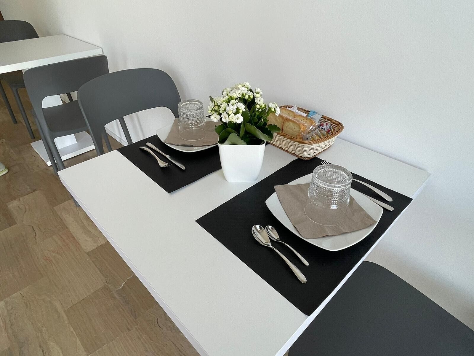 A dining table set with black placemats, silverware, a clear glass, a white vase with flowers, and a basket with bread and condiments.