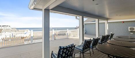 A covered outdoor deck with a view of the beach, featuring a long table and chairs, and a brick wall with an air conditioning unit.