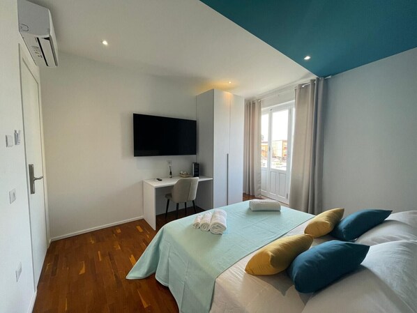 Habitació d'hotel amb llit, escriptori, televisió i finestra amb cortines.