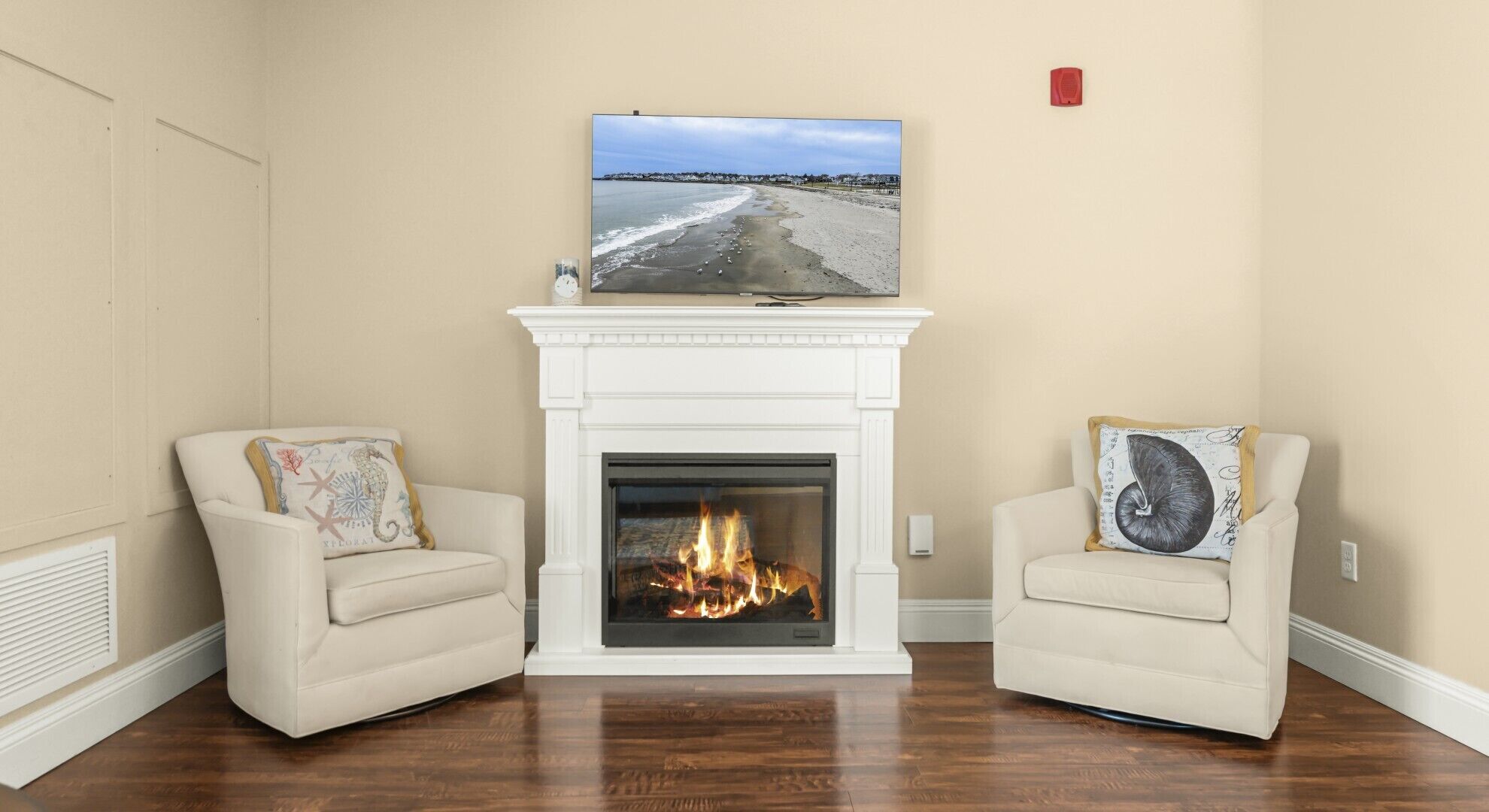 Un salon avec une cheminée, deux fauteuils, un tableau encadré représentant une plage et un détecteur d’incendie rouge fixé au mur.