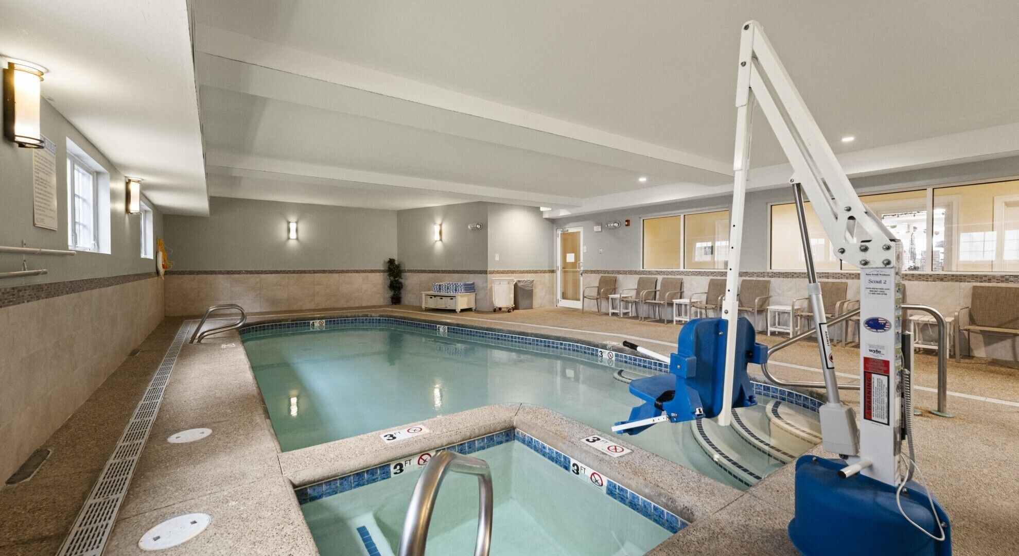 An indoor swimming pool with a lifeguard chair, a pool ladder, and a seating area with chairs.