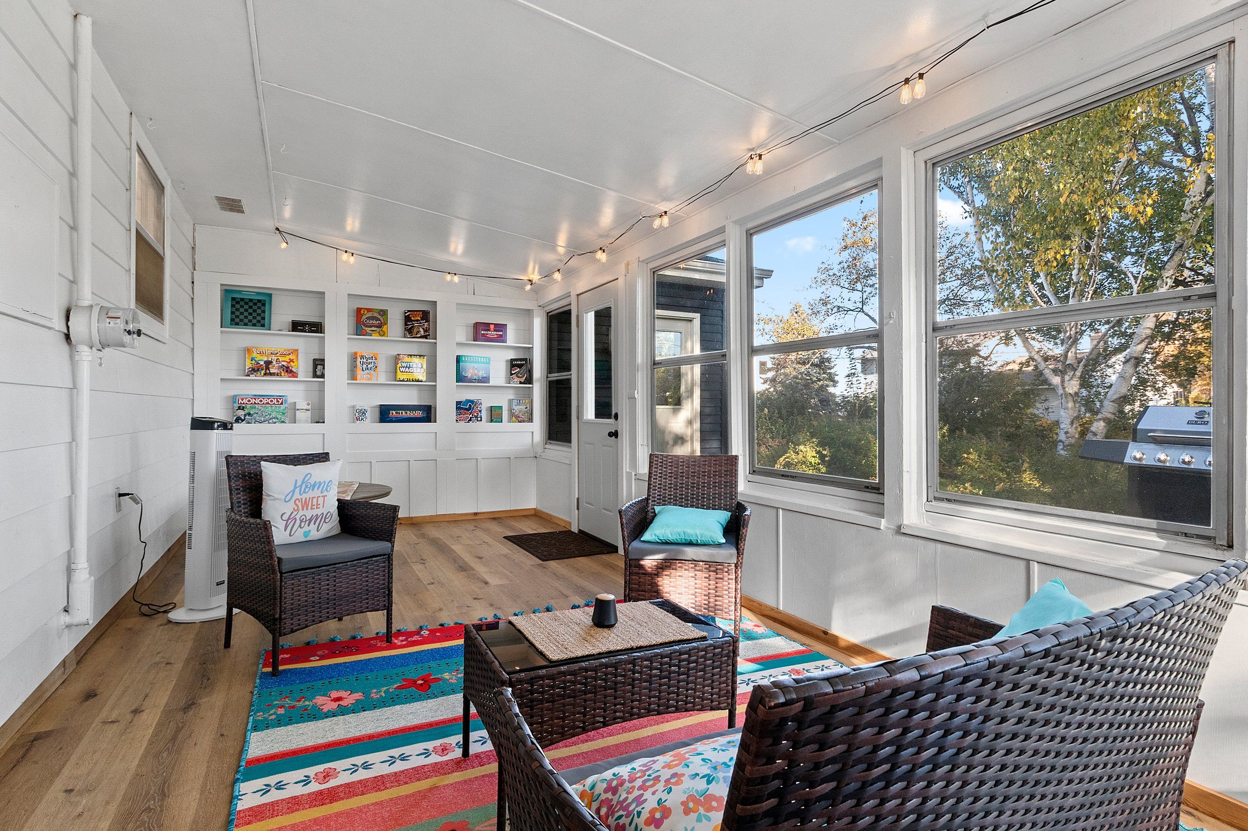 A sunlit room with a large window, wicker furniture, a colorful rug, and a bookshelf with various books.