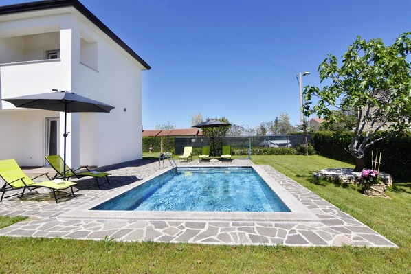 A swimming pool with lounge chairs and a patio umbrella, surrounded by a stone deck and a well-maintained lawn. - Relaxing Holiday Home With Shared Pool (Umag)