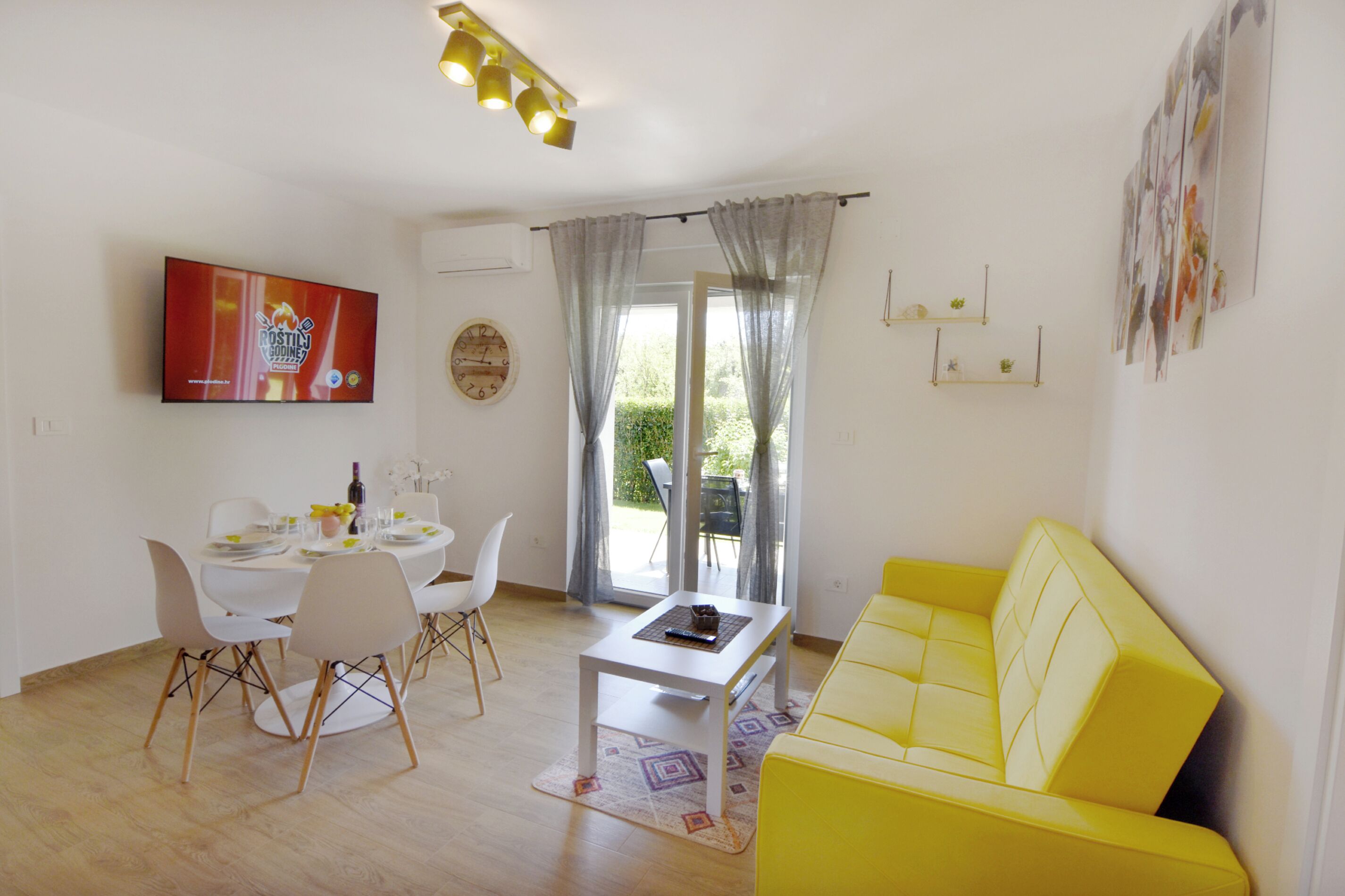 A modern living room with a yellow sofa, a dining table with chairs, a coffee table, and a TV mounted on the wall.