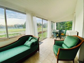 Una stanza con un divano verde, una poltrona verde, un tavolo da pranzo con sedie e ampie finestre che si affacciano su un paesaggio verdeggiante.