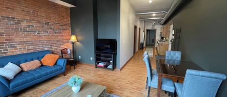 Una sala de estar moderna con un sofá azul, una mesa de centro y sillas azules. Se ve una pared de ladrillo, una chimenea y un comedor con una mesa y sillas.