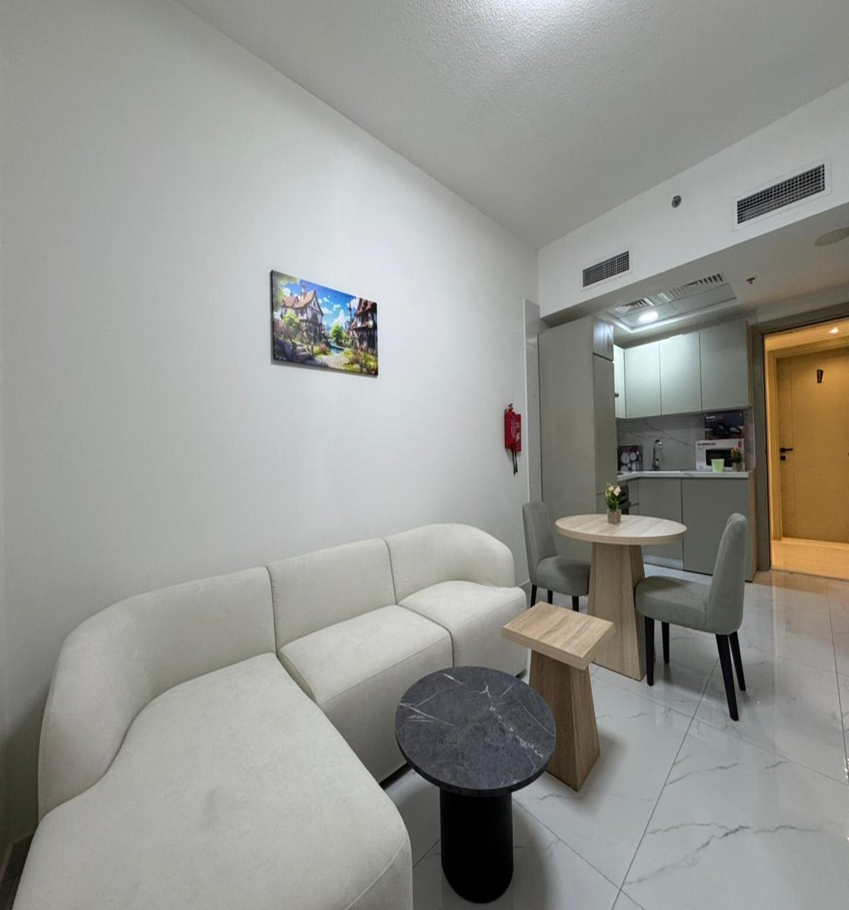 Un salon moderne avec un canapé, une table ronde et une petite table basse. Un tableau orne le mur, et on aperçoit un coin cuisine au fond.
