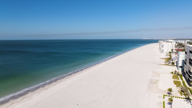 A beach with a clear view of the ocean, a sandy shore, and a row of modern buildings with balconies. - Charming 1/1 home across the street from the ocean sunsets, dock space included! (Bonita Springs)