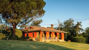 A red house with a porch, surrounded by trees and a well-maintained lawn. - Panoramic Tuscan villa with infinity pool, exclusive use, private garden (Montecarlo)