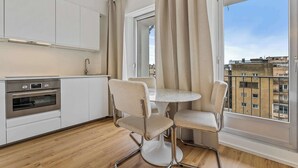 Een moderne keuken met witte kasten en roestvrijstalen apparatuur, een ronde eettafel met twee stoelen en een groot raam met gordijnen.