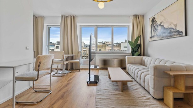 Een moderne woonkamer met een bank, salontafel, stoel, eettafel en een groot raam met uitzicht op gebouwen.