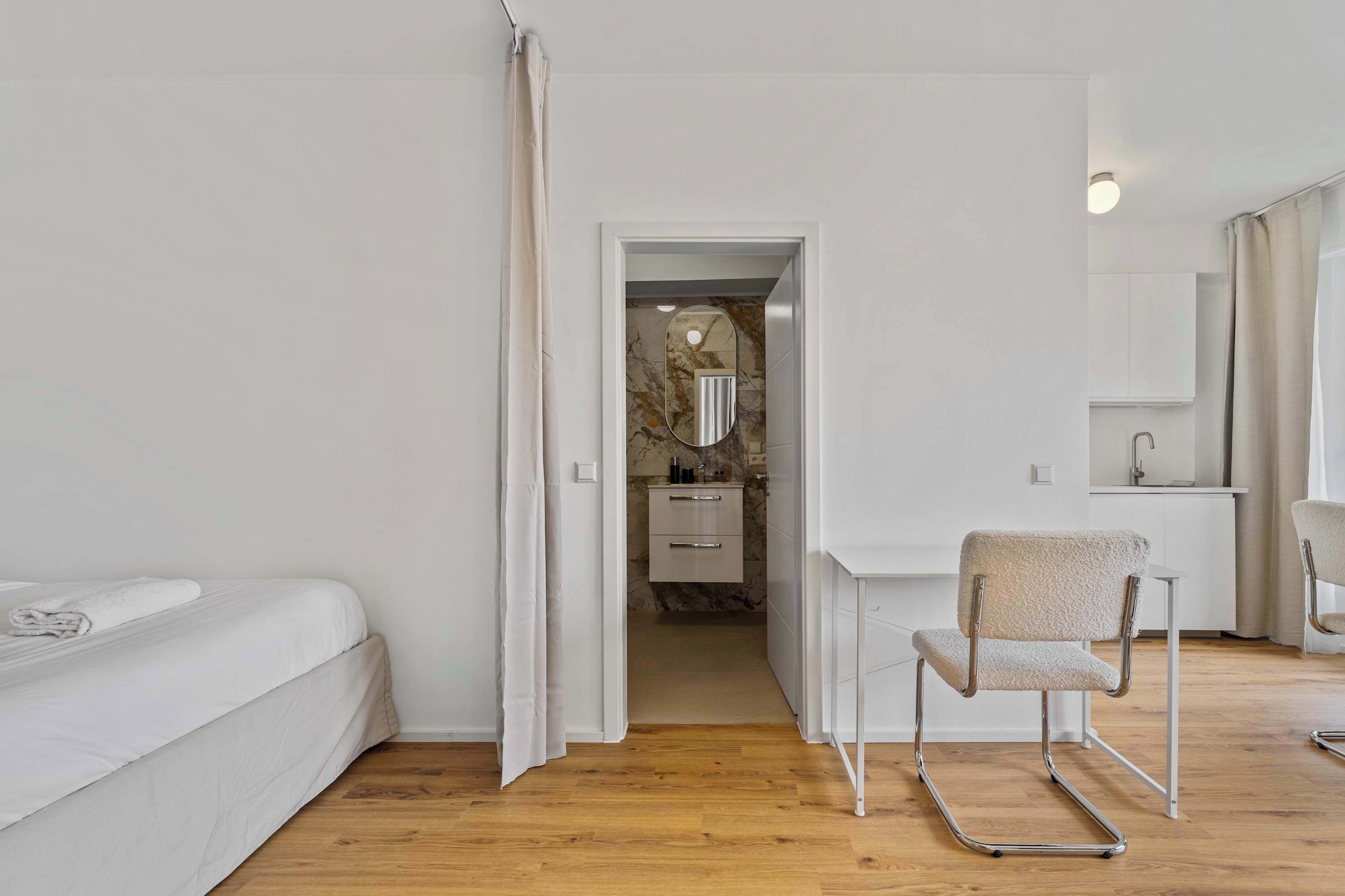 Une chambre avec un plancher en bois, un lit blanc, une chaise et une salle de bain visible à travers une porte ouverte.